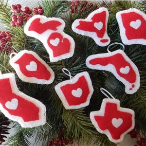 May include: Nine red felt ornaments shaped like different states in the United States. Each ornament has a white heart in the center.