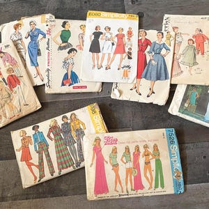 May include: A collection of vintage Simplicity sewing patterns. The patterns feature illustrations of dresses, pantsuits, and other garments. The covers display various colors, including blue, red, green, and pink. The patterns are arranged on a dark wooden surface.