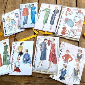 May include: A collection of seven vintage Simplicity printed sewing pattern envelopes featuring illustrations of dresses, tops, and baby clothing, accompanied by a yellow measuring tape. Each envelope displays garment sketches, prices, and pattern numbers.