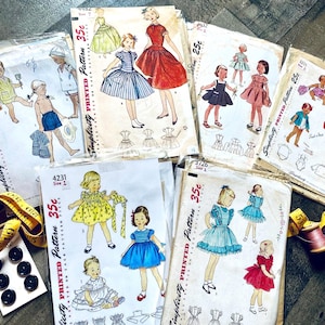 May include: A collection of vintage Simplicity sewing patterns for children's clothing. The patterns feature illustrations of dresses, shorts, and jackets. Accessories include a measuring tape, buttons, and thread spools. The patterns are priced at 25¢ and 35¢.