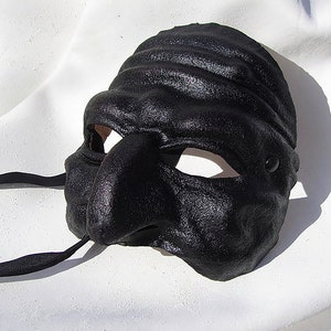 May include: A black leather mask with a textured finish. The mask has a large nose, eye openings, and a strap. This mask is likely for costume or theatrical purposes.