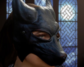 May include: A black leather mask with a pointed snout and eye holes. The mask is worn by a person with dark hair, and the background is a blurry image of stained glass windows.