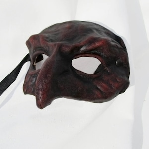 Könnte beinhalten: Eine dunkelbraune Leder-Maskenballmaske mit einer langen, spitzen Nase und einem einzigen Augenloch. Die Maske hat ein schwarzes Lederband.