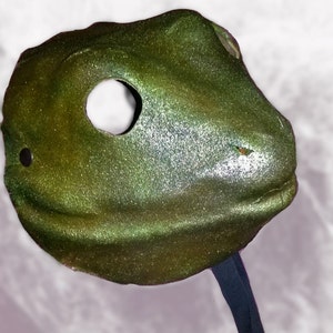 May include: A metallic green mask with a single eye hole and a black strap. The mask has a textured surface and a slightly curved shape, suggesting a stylized animal or creature design. The mask is likely for costume or decorative purposes.