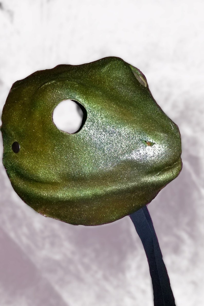 May include: A metallic green mask with a single eye hole and a black strap. The mask has a textured surface and a slightly curved shape, suggesting a stylized animal or creature design. The mask is likely for costume or decorative purposes.