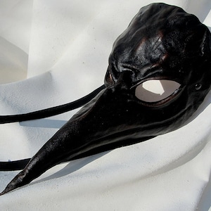 May include: A black leather masquerade mask with a long, pointed nose and a single eye hole. The mask has a textured surface and is tied with black ribbons.