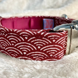 Pu&ograve; includere: Un collare per cani con un motivo a onde rosse e bianche. Il collare ha una fibbia in metallo argentato e un interno magenta. Il motivo a onde &egrave; un disegno ripetitivo di linee curve bianche su sfondo rosso.