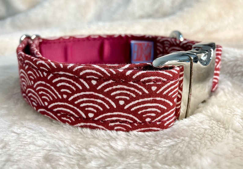 Pu&ograve; includere: Un collare per cani con un motivo a onde rosse e bianche. Il collare ha una fibbia in metallo argentato e un interno magenta. Il motivo a onde &egrave; un disegno ripetitivo di linee curve bianche su sfondo rosso.