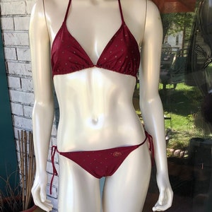 May include: A burgundy bikini set on a mannequin. The halter-top and low-rise bottoms are adorned with small, gold-colored star-shaped accents. The bottoms feature a gold logo. The mannequin is displayed outdoors.