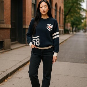 May include: Navy blue sweater with white accents, featuring a shield emblem and the number "58". The sweater has white stripes on the sleeves and a cream-colored hem and cuffs. The person is wearing black jeans.