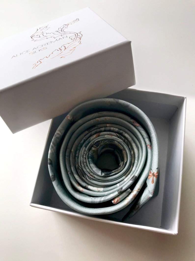 May include: A rolled up green silk necktie with a floral pattern. The necktie is in a white box with the words 'Alice Ackerman Silks' printed on the lid.