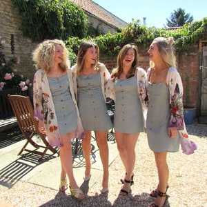 May include: Four women wearing light green button-down dresses and floral print kimonos. They are standing outside on a sunny day. The dresses have a simple, classic design, and the kimonos add a pop of color and pattern.