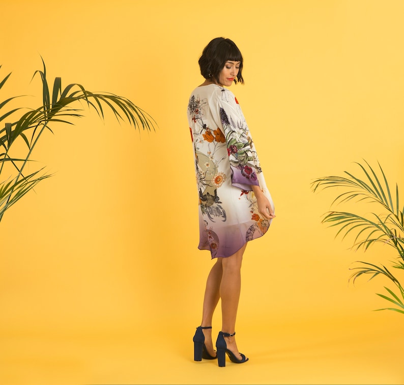 May include: A flowing, knee-length dress with a floral and bird print in shades of cream, orange, and purple. The dress has long sleeves and a slight ombre effect at the hem. The model is wearing navy blue heeled sandals.