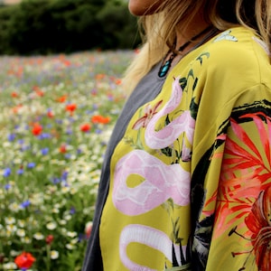 Op de afbeelding: Een gele kimono-achtige top met een roze slangenontwerp en bloemaccenten. De top is gelaagd over een grijs shirt. De achtergrond toont een veld met wilde bloemen.
