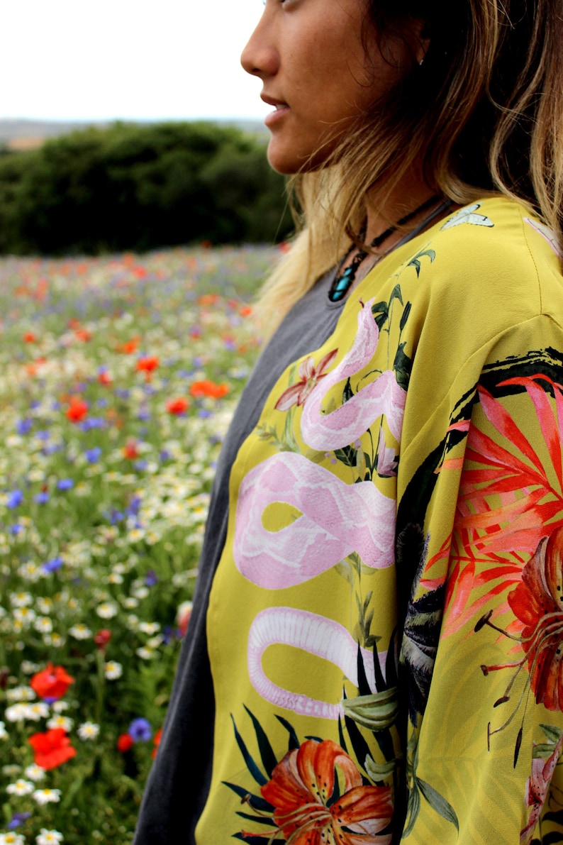 Op de afbeelding: Een gele kimono-achtige top met een roze slangenontwerp en bloemaccenten. De top is gelaagd over een grijs shirt. De achtergrond toont een veld met wilde bloemen.