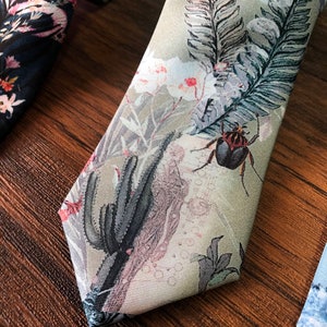 May include: A green silk tie with a floral pattern. The pattern includes pink flowers, green leaves, and a black beetle. The tie is on a wooden surface.
