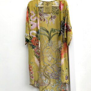 Op de afbeelding: Een lange, open kimono in een lichtgele tint, versierd met een levendige botanische print. Het ontwerp toont orchidee&euml;n, slangen, vlinders en andere flora en fauna. Een label met tekst hangt aan de hanger.