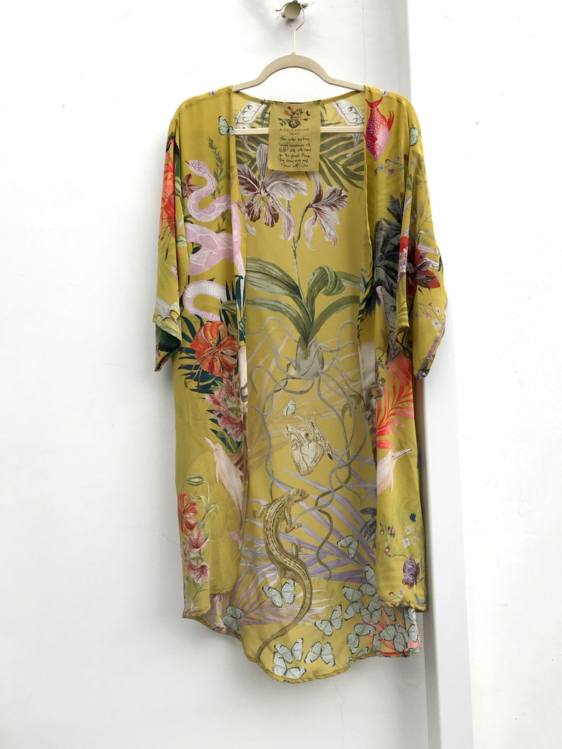 Op de afbeelding: Een lange, open kimono in een lichtgele tint, versierd met een levendige botanische print. Het ontwerp toont orchidee&euml;n, slangen, vlinders en andere flora en fauna. Een label met tekst hangt aan de hanger.