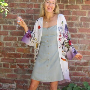 May include: A light green button-down dress paired with a floral print kimono. The kimono features a white base with colorful flowers and a purple ombre effect on the sleeves. The background is a red brick wall.