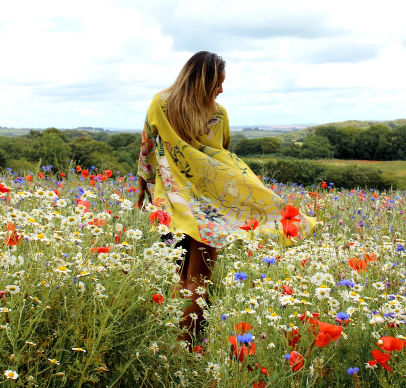 Op de afbeelding: Een gele kimono met een bloemenpatroon wordt gedragen in een veld met wilde bloemen. De stof van de kimono wappert in de wind. Het veld is gevuld met witte madeliefjes, rode klaprozen en blauwe korenbloemen, wat een levendige sc&egrave;ne cre&euml;ert.