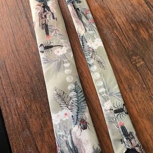 May include: Two green silk ties with a floral pattern. The pattern includes cacti, flowers, and insects. The ties are on a wood surface.