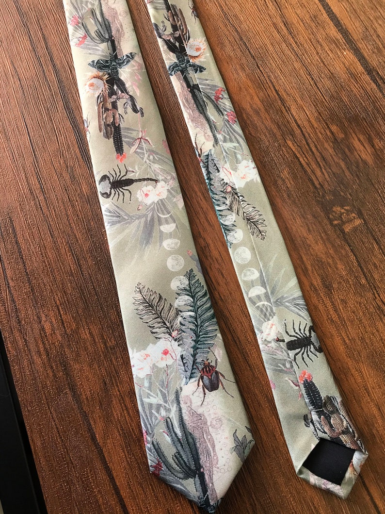May include: Two green silk ties with a floral pattern. The pattern includes cacti, flowers, and insects. The ties are on a wood surface.