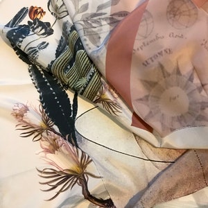 K&ouml;nnte beinhalten: Ein wei&szlig;er Seidenschal mit einem Blumendruck, der einen Kaktus, eine Blume und einen Zweig zeigt. Der Schal hat auf der R&uuml;ckseite ein pinkfarbenes und wei&szlig;es geometrisches Muster. Der Text "September, dont..." ist auf der R&uuml;ckseite des Schals sichtbar.