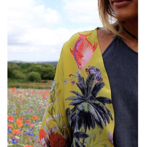 Op de afbeelding: Een gele kimono met een tropische vis- en palmboomprint. De kimono heeft een levendig ontwerp met rode, paarse en groene accenten. De achtergrond toont een veld met wilde bloemen onder een bewolkte hemel.
