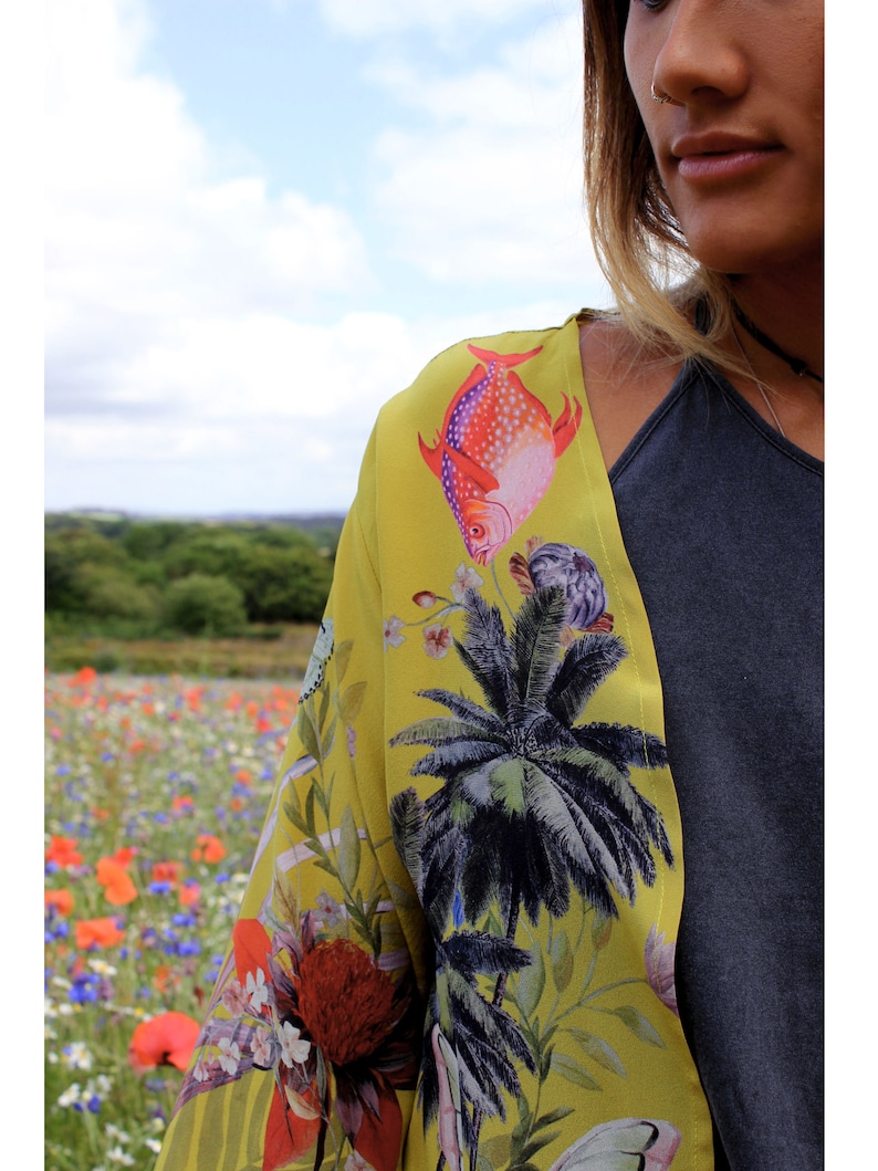 Op de afbeelding: Een gele kimono met een tropische vis- en palmboomprint. De kimono heeft een levendig ontwerp met rode, paarse en groene accenten. De achtergrond toont een veld met wilde bloemen onder een bewolkte hemel.