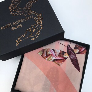 K&ouml;nnte beinhalten: Ein rosa Seidentuch mit einem braunen und gr&uuml;nen Schmetterlingsmuster. Das Tuch befindet sich in einer schwarzen Schachtel mit dem Text "Alice Acreman Silks" auf dem Deckel.