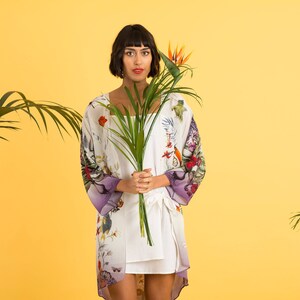 May include: A woman wearing a white kimono-style robe with a floral and bird print, accented with purple sleeves. She holds a green plant and stands against a yellow background. The robe is paired with a white dress.