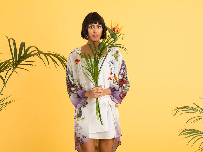 May include: A woman wearing a white kimono-style robe with a floral and bird print, accented with purple sleeves. She holds a green plant and stands against a yellow background. The robe is paired with a white dress.