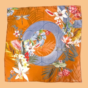 Könnte beinhalten: Ein quadratisches orangefarbenes Seidentuch mit Blumen- und Libellenmuster. Der Schal hat einen großen blauen Kreis mit einem Blumenarrangement aus weißen, roten und blauen Blumen. Zwei Libellen sind sichtbar.