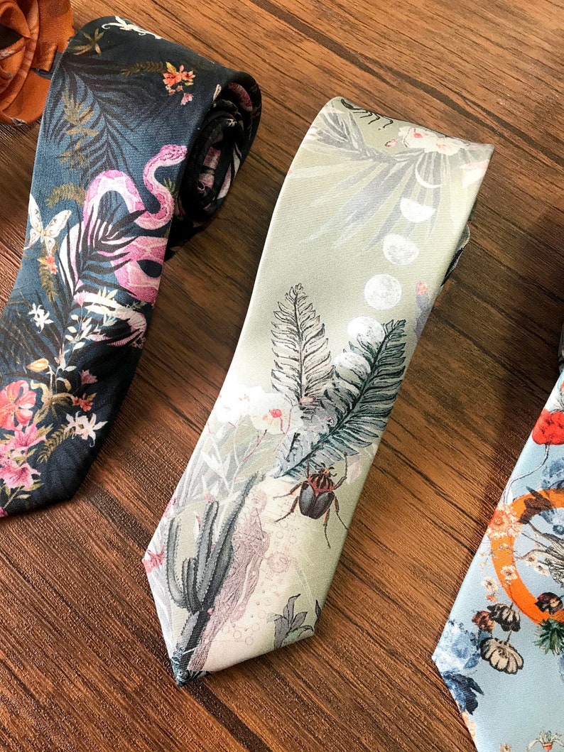 May include: Three patterned neckties, one with a dark blue background with pink flamingos and green plants, one with a light green background with white moons, gray ferns, and a beetle, and one with a light blue background with orange and blue flowers and a butterfly.