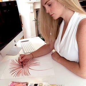 Op de afbeelding: Een persoon schildert een aquarelillustratie van een gestileerde, roodbruine palmblad op wit papier. De kunstenaar gebruikt een penseel en een palet met waterverf. Een computer en een toetsenbord zijn zichtbaar op de achtergrond.