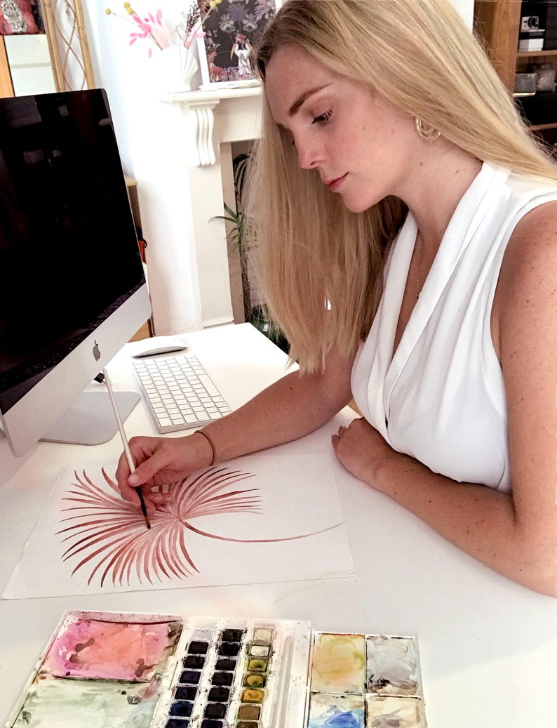 Op de afbeelding: Een persoon schildert een aquarelillustratie van een gestileerde, roodbruine palmblad op wit papier. De kunstenaar gebruikt een penseel en een palet met waterverf. Een computer en een toetsenbord zijn zichtbaar op de achtergrond.