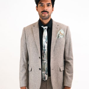 May include: A man wearing a gray suit with a black shirt and a patterned green tie. He is also wearing a green pocket square.