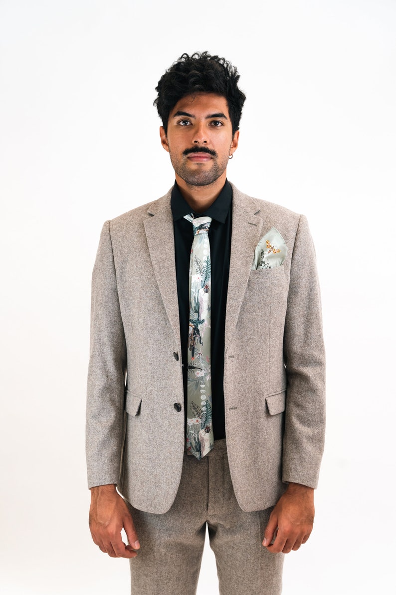 May include: A man wearing a gray suit with a black shirt and a patterned green tie. He is also wearing a green pocket square.
