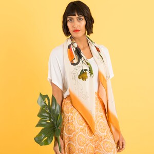 May include: A woman wearing a white t-shirt and orange and white patterned pants with a white scarf featuring a toucan and floral design. She is holding a large green leaf.