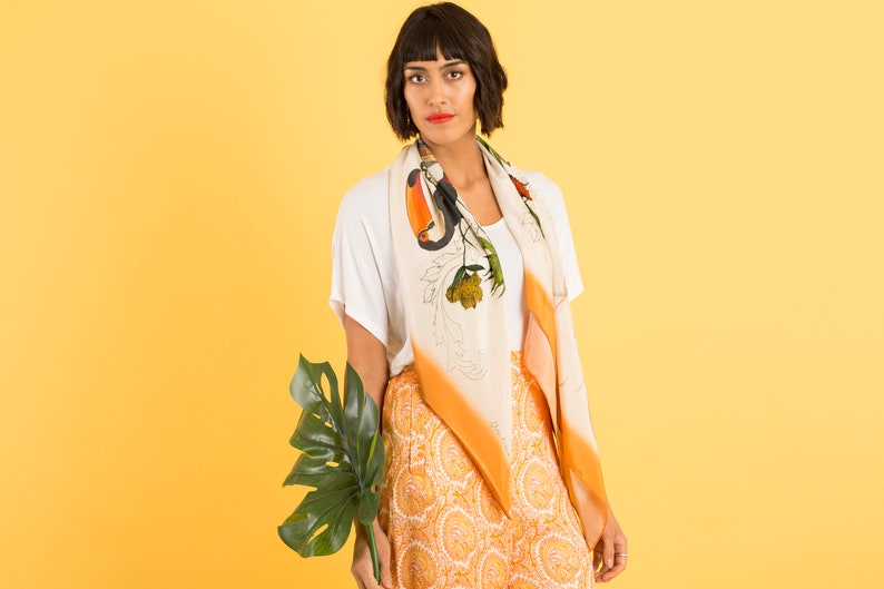 May include: A woman wearing a white t-shirt and orange and white patterned pants with a white scarf featuring a toucan and floral design. She is holding a large green leaf.