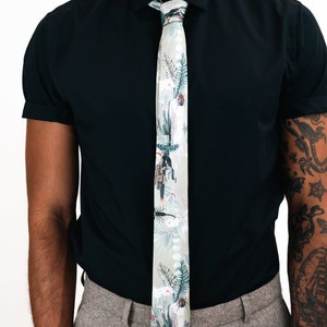 May include: A light gray tie with a floral print featuring cacti, flowers, and insects. The tie is tied around a black collared shirt.