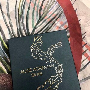K&ouml;nnte beinhalten: Eine schwarze Box mit goldener Beschriftung, die "Alice Acreman Silks" lautet. Die Box ist mit einem goldenen floralen Design verziert. Die Box steht auf einem beige-roten gemusterten Stoff.
