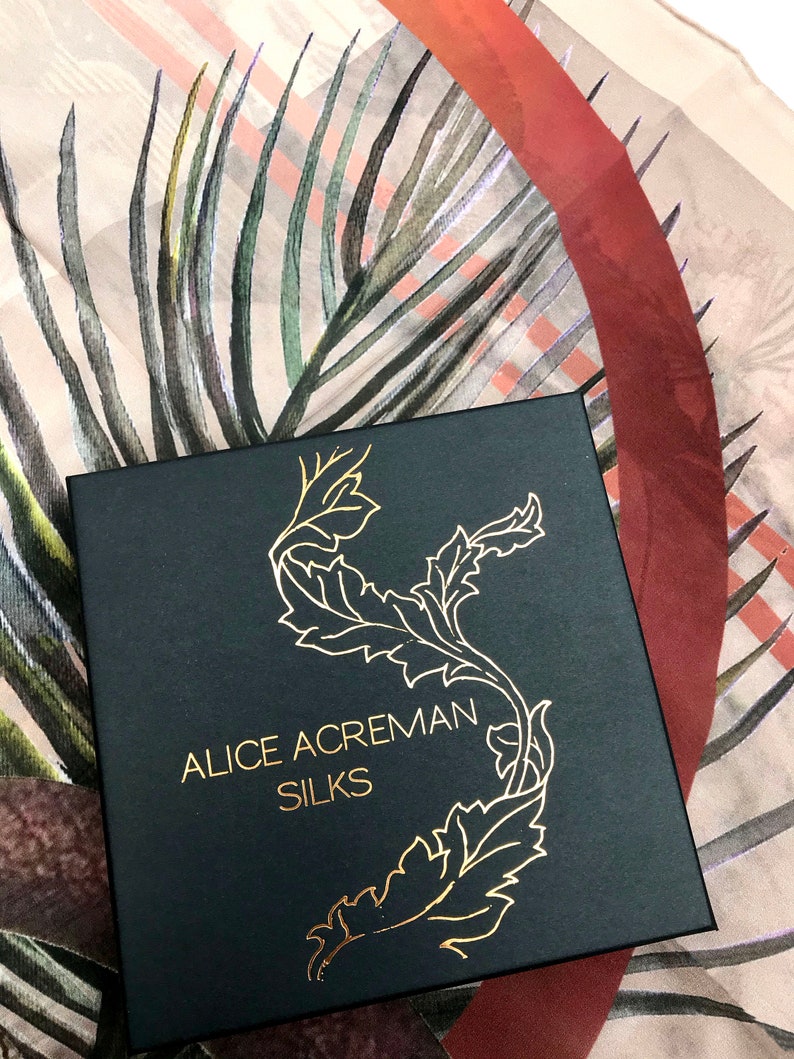 K&ouml;nnte beinhalten: Eine schwarze Box mit goldener Beschriftung, die "Alice Acreman Silks" lautet. Die Box ist mit einem goldenen floralen Design verziert. Die Box steht auf einem beige-roten gemusterten Stoff.