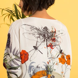 May include: White, long-sleeved top with a botanical and dragonfly print. The design features red, orange, and white flowers, along with green foliage. The background is a bright yellow.