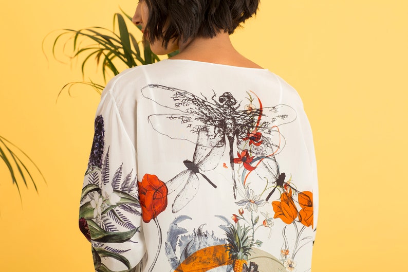 May include: White, long-sleeved top with a botanical and dragonfly print. The design features red, orange, and white flowers, along with green foliage. The background is a bright yellow.