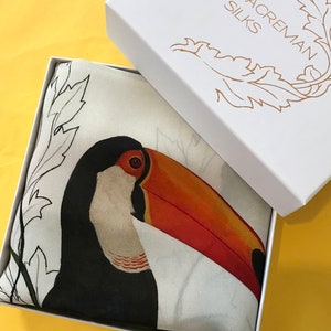 May include: A white box with the text "Alice Acraman Silks" printed on the top. The box is open and contains a white silk scarf with a black and orange toucan illustration.