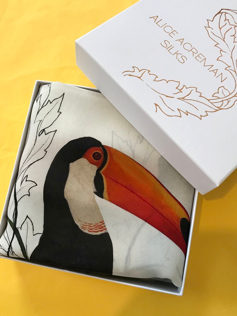 May include: A white box with the text "Alice Acraman Silks" printed on the top. The box is open and contains a white silk scarf with a black and orange toucan illustration.