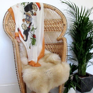 May include: A white wicker peacock chair with a patterned orange and white scarf draped over the back. The chair has a fluffy white sheepskin rug on the seat. A potted green plant is in the background.