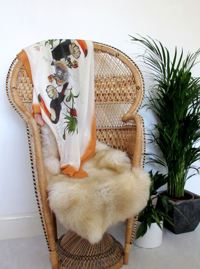 May include: A white wicker peacock chair with a patterned orange and white scarf draped over the back. The chair has a fluffy white sheepskin rug on the seat. A potted green plant is in the background.