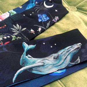Könnte beinhalten: Drei dunkle Stoff-Haarbänder mit einzigartigen Designs. Eines zeigt einen Blauwal, der in einem sternenklaren Nachthimmel schwimmt. Ein anderes hat ein Blumenmuster mit rosa und grünen Akzenten. Das dritte Haarband zeigt einen Mond und Blumen.